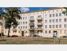 Mehrstöckiges Wohngebäude im Stil der Nachkriegsarchitektur mit heller Fassade und mehreren Balkonen. Im Vordergrund befindet sich eine Grünfläche mit einem Baum, Wäscheleinen und geparkten Autos.