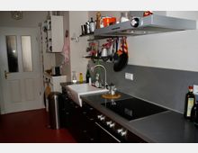 Kleine moderne Küche mit dunklen Schränken, einem weißen Keramikspülbecken und einer grauen Arbeitsplatte. An der Wand hängen Küchenutensilien, während eine Herdplatte und ein Dunstabzug auf der rechten Seite sichtbar sind; links befindet sich eine Tür mit Milchglasfenstern.