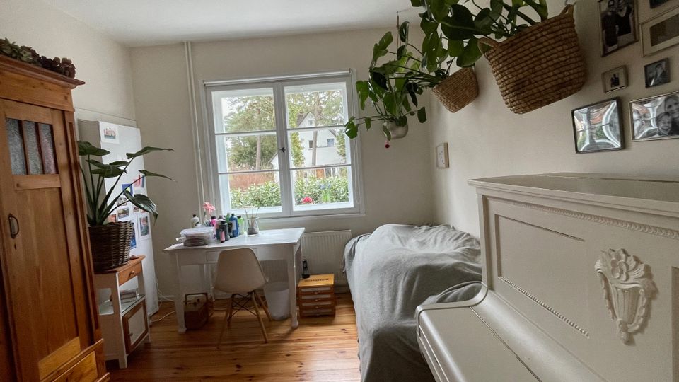 Helles Schlafzimmer mit Holzdielen, einem Bett an der rechten Wand und einem großen Fenster am Ende des Raums mit Schreibtisch davor. Links stehen ein Holzschrank und ein Kühlschrank, rechts hängen Pflanzenkörbe und mehrere gerahmte Fotos an der Wand.