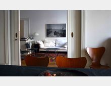 Wohnzimmeransicht mit einer weißen Couch, einem runden Couchtisch und einem schwarz-weißen Wandbild. Im Vordergrund sind Holzstühle und ein Tisch zu sehen, während links ein Bücherregal mit einer Tischlampe steht.