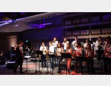 Eine Musikgruppe aus jungen Musikerinnen und Musikern spielt auf einer Bühne in einem Innenraum, umgeben von indirekter lila Beleuchtung. Im Vordergrund sind Saxophonisten und Trompeter zu sehen, während im Hintergrund ein Segelflugzeug von der Decke hängt.