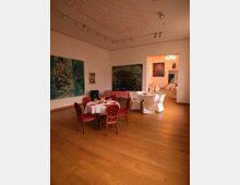 Ein eleganter Innenraum mit Holzboden, runden Tischen mit weißer Tischdecke und dekorierten Stühlen in Rot und Gold. An den Wänden hängen großformatige Kunstwerke, und die Decke ist mit einem leuchtenden Netz aus kleinen Lichtern verziert.