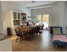 Heller Wohn- und Essbereich mit Holzboden, großem Holzesstisch mit mehreren Stühlen und einem weißen Vitrinenschrank an der Wand. Eine breite Glasschiebetür führt auf eine verschneite Terrasse bzw. in den Garten; rechts steht ein blaues Sofa mit Kissen und Decke.