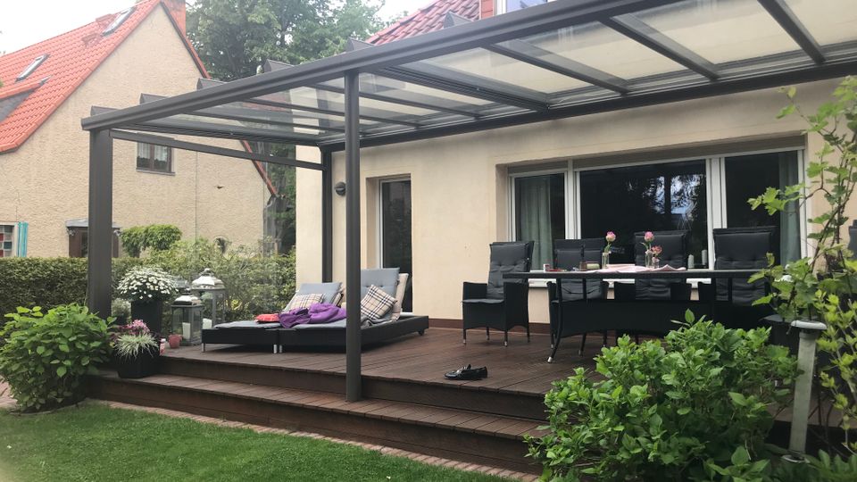 Überdachte Holzterrasse an einem Wohnhaus mit großem Glasdach und Metallstützen, davor Rasen und Gartensträucher. Unter der Überdachung stehen Liege, Sitzgruppe und Esstisch vor einer breiten Schiebetür zur Hausfassade.