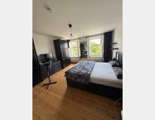 Helles Schlafzimmer mit Holzdielenboden, großem Bett und schwarzen Möbeln wie Kleiderschrank und Kommode mit Fernseher. Zwei große Fenster mit dunklen Vorhängen und Heizkörpern darunter lassen viel Tageslicht herein; an der Decke hängt eine geometrische Pendelleuchte.