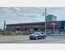 Straßenszene mit dem Bahnhof "Warschauer Straße" in Berlin im Hintergrund, einem modernen Backsteingebäude mit Glasfassaden und Graffiti. Im Vordergrund ein Auto auf der Straße, ein Fußgänger auf dem Bürgersteig und eine Kirchturmspitze links im Bild.