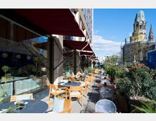 Außenbereich eines Cafés mit Holztischen und Stühlen sowie großen Pflanzenkübeln entlang eines Gehwegs. Im Hintergrund ist die Kaiser-Wilhelm-Gedächtniskirche und ein moderner Straßenbereich zu sehen.