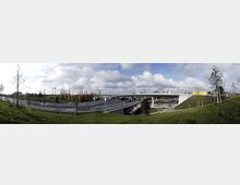 Weitwinkelaufnahme einer mehrspurigen Autobahn mit einer modernen Überführung, umgeben von grünen Wiesen und jungen Bäumen. Im Hintergrund sind Industriegebäude und ein bewölkter Himmel zu sehen.