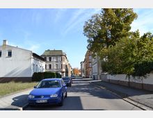 Straßenansicht in einer Wohngegend mit gepflastertem Gehweg, geparkten Autos, mehreren Bäumen und Gebäuden im Hintergrund, darunter ein älteres Haus mit grüner Dachdeckung und rote Backsteinfassaden. Der Himmel ist klar und blau.