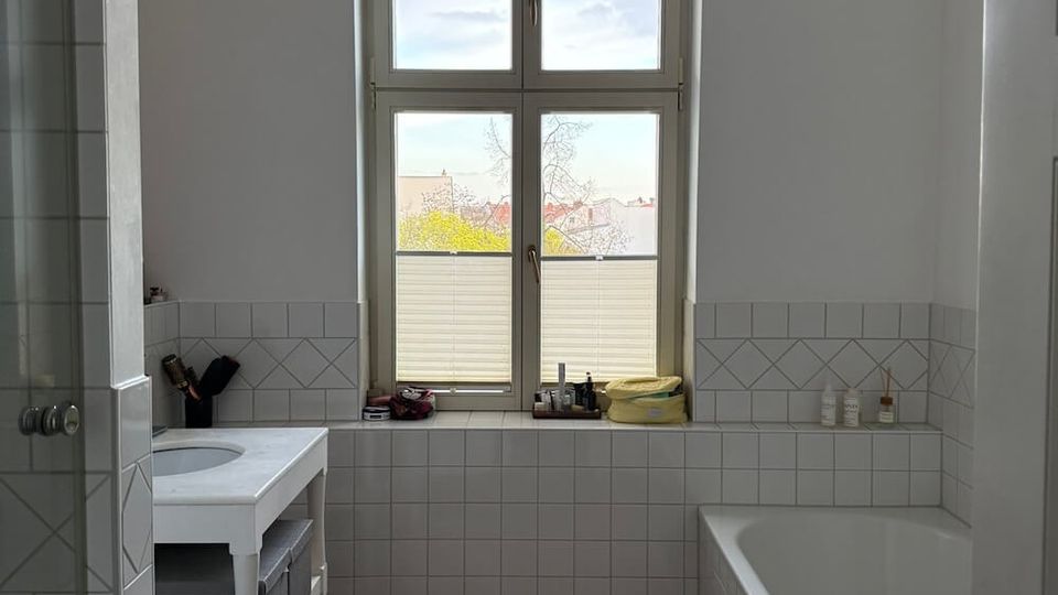 Bright bathroom with white half-height wall tiles and a wood floor, featuring a large arched window at the far end. A bathtub runs along the right wall, a small vanity sink sits on the left, and a glass shower partition is visible near the entrance.