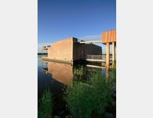 Holzgebäude auf einer Wasserfläche, mit moderner, rechteckiger Architektur und horizontalen Holzlatten verkleidet; eine metallene Fußgängerbrücke verbindet das Gebäude mit einer angrenzenden Struktur. Im Vordergrund wachsen Schilf und andere Pflanzen am Ufer.