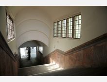 Treppenhaus eines historischen Gebäudes mit gewölbter Decke, bleiverglasten Fenstern und Wandverkleidung aus Stein. Unten führt die Treppe zu einem breiten Durchgang mit Blick ins Freie.