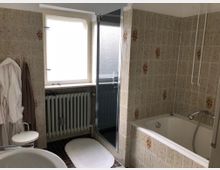 Badezimmer mit beige-gemusterten Fliesenwänden, Badewanne und Dusche mit Glastür. Ein Fenster mit Milchglas befindet sich über einem Heizkörper, daneben hängen zwei Bademäntel an der Wand.