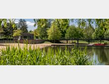 Uferbereich mit kleinem Sandstrand und einem modernen Gebäude aus Backstein, umgeben von Bäumen. Im Vordergrund Schilfgras und ein ruhiger See mit einem Steg, an dem ein rotes Kajak liegt.