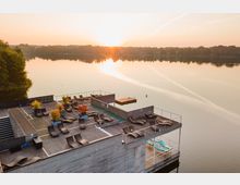 Terrasse eines modernen Holzgebäudes auf einem See, mit Liegestühlen, Pflanzen in großen Kübeln und einem kleinen Schwimmbecken; im Hintergrund spiegelglattes Wasser und ein Sonnenuntergang, umrahmt von Bäumen.