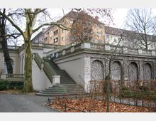 Steinernes Treppenhaus mit Balustrade in einer parkähnlichen Umgebung, flankiert von kahlen Bäumen und rankenbewachsenen Wänden; im Hintergrund mehrstöckige Wohngebäude. Der Boden ist mit Herbstlaub bedeckt.