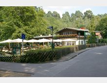 Ein traditionelles Gasthaus mit Holzfassade, umgeben von Bäumen und grüner Landschaft. Vor dem Gebäude befinden sich eine Terrasse mit weißen Sonnenschirmen, Sitzplätzen für Gäste sowie ein Holzzaun entlang des Weges.