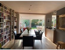 Esszimmer mit einem langen Holztisch und sechs schwarzen Stühlen, flankiert von einer großen Bücherwand auf der linken Seite. Im Hintergrund befindet sich eine breite Glasfront mit Blick auf einen grünen Garten mit Gartenmöbeln.