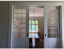 Weiß lackierte Schiebetüren mit verzierten Glasfenstern führen in einen Essbereich mit einem langen Holztisch und mehreren Stühlen. Im Hintergrund ist ein großes Fenster erkennbar, das Tageslicht hereinlässt.