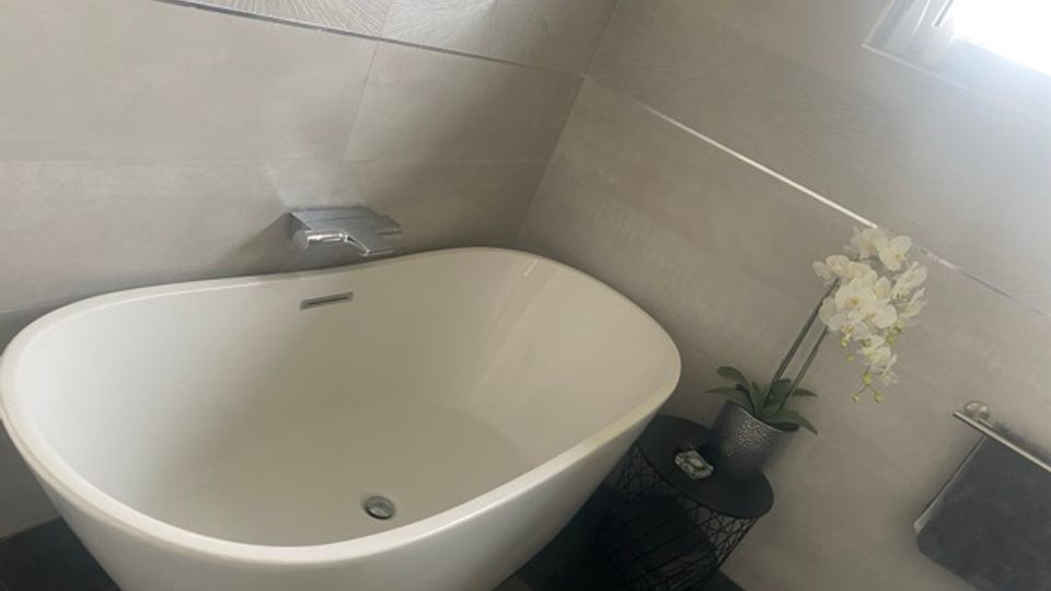 Modernes Badezimmer mit freistehender weißer Badewanne vor grauen Wandfliesen und dunklem Boden, ergänzt durch eine kleine Ablage mit Pflanze. Ein Fenster sorgt für Tageslicht, daneben sind ein Handtuchhalter und Armaturen an der Wand zu sehen.