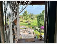 Blick aus einer Tür auf eine gepflasterte Terrasse mit Pflanzen und Sitzmöbeln, die in einen weitläufigen Garten mit Rasenflächen, Blumen und Bäumen übergeht. Im Vordergrund hängt ein Vorhang aus Perlenketten, und eine Metalltreppe führt nach draußen.