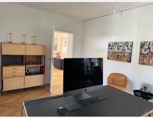 Ein modernes Arbeitszimmer mit einem großen Monitor auf einem schwarzen Schreibtisch. Im Hintergrund befinden sich ein Holzregal mit Dekorationen, eine offene Tür, die in einen weiteren Raum führt, sowie zwei abstrakte Gemälde an der weißen Wand. Auf dem Boden liegt ein Fischgrätparkett.