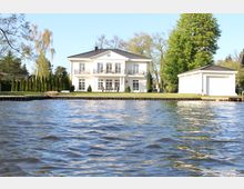 Weiße Villa im klassischen Stil mit symmetrischen Fenstern, Balkonen und einem Satteldach, gelegen an einem Gewässer. Daneben befindet sich eine Garage, umgeben von gepflegtem Rasen, Bäumen und einer Hecke.