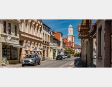 Blick entlang einer schmalen Altstadtstraße mit Kopfsteinpflaster, historischen Fassaden und kleinen Geschäften mit Ausleger-Schildern. Am Straßenende ragt ein hoher Kirchturm mit grüner Kuppel über die Dächer.