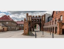 Historisches Stadttor aus rotem Backstein mit Zinnen und Bögen, flankiert von Gebäuden mit roten Ziegeldächern; rechts eine kleine mittelalterliche Mauer mit Arkaden. Im Hintergrund ist eine gepflasterte Straße und modernes Gebäude zu sehen.