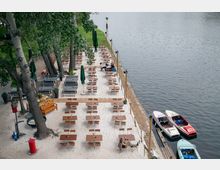 Ein Biergarten mit Holztischen und Stühlen auf hellem Sandboden, umgeben von Bäumen, direkt an einem Flussufer. Am Ufer sind mehrere kleine Boote festgemacht.