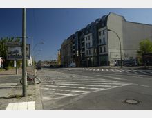 Straßenkreuzung mit Zebrastreifen in einem urbanen Bereich, flankiert von mehrstöckigen Gebäuden, darunter modernisierte Wohn- und Geschäftsgebäude. Im Vordergrund stehen ein Fahrrad an einem Laternenmast und ein Werbeschild mit Straßenname „Liesenstraße“, während am Horizont weitere Architektur und Fahrzeuge sichtbar sind.