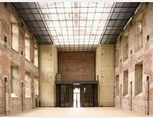 Innenraum eines historischen Gebäudes mit freigelegten Backsteinwänden, hohen Fensternischen und einem modernen Glasdach. Der Boden ist aus hellem Material, und im Hintergrund befindet sich eine gläserne Eingangstür mit einem erhöhten Balkonbereich darüber.