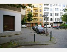 Straßenecke in einer Wohngegend mit gepflastertem Gehweg und parkenden Autos entlang der Straße. Im Hintergrund befinden sich mehrstöckige Wohnhäuser mit Balkonen sowie ein Straßenschild und eine Uhr auf einem Pfeiler.