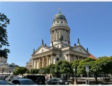 Historisches Gebäude mit Kuppel: Das Bild zeigt den Deutschen Dom in Berlin, ein klassizistisches Bauwerk mit Säulen, Figurenschmuck und einer großen, grünen Kuppel. Im Vordergrund ist eine Reihe von Bäumen, geparkte Autos und ein Verkehrszeichen sichtbar.