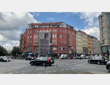 Straßenkreuzung in einer Stadt mit mehrspurigen Straßen und Straßenbahnschienen, umgeben von mehrstöckigen Gebäuden, darunter ein auffälliges rotes Eckhaus mit Gerüst an der Fassade. Im Vordergrund fahren Autos, und Passanten sind auf dem Gehweg sichtbar.