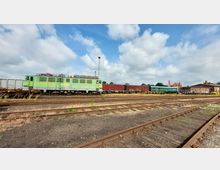 Güterbahnhof mit mehreren Gleisen und abgestellten Zügen, darunter eine grün-gelbe Lokomotive mit der Aufschrift „EGP“. Im Hintergrund befinden sich ein langes Backsteingebäude und wolkiger Himmel.