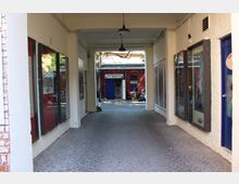 Überdachter Durchgang mit weiß gestrichenen Wänden, Kopfsteinpflasterboden und hängenden Lampen, an den Seiten mit großen verglasten Schaukästen und Türen. Am Ende öffnet sich der Gang zu einem Innenhof mit einem roten Backsteingebäude und blauem Eingang.