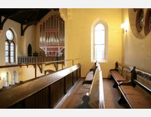 Innenraum einer Kirche mit holzgetäfelten Sitzbänken auf einer Empore. Im Hintergrund ist eine große Orgel vor einer Wand mit roten und weißen Verzierungen sowie mehrere farbige Glasfenster sichtbar.