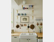 Küche mit weißen Fliesen im Hintergrund, offener weißer Spüle und Arbeitsplatten aus Holz. Über der Spüle befinden sich drei offene Regale mit Geschirr, Gläsern und Dekorationsgegenständen, sowie eine Messing-Stange, an der Kupfertöpfe und Küchenutensilien hängen. Rechts sind Teile eines traditionellen Kachelherdes sichtbar.