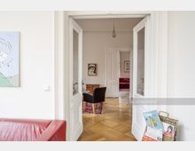 Ein heller Raum mit weißen Wänden und doppelflügeligen Glastüren, die in weitere Räume mit Holzboden und klassischen weißen Holztüren führen. Im Vordergrund steht ein rotes Ledersofa, ein moderner Zeitschriftenhalter, und an der Wand hängt ein Gemälde.