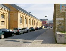 Straßenszene in einem Industrie- oder Gewerbegebiet mit gelben Backsteingebäuden aus mehreren Fensterreihen. Geparkte Autos stehen entlang einer gepflasterten Straße, und an einer Gebäudewand ist ein Schild mit der Aufschrift „Reithalle B“ angebracht.