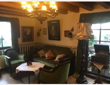 Gemütliches Wohnzimmer im Vintage-Stil mit alten Möbeln, darunter grüne Polstersessel und ein Sofa, ein runder Holztisch mit Spitzendecke sowie eine Stehlampe mit verziertem Schirm. Holzdecke, Wände mit floralen Ornamenten dekoriert, und ein großes Fenster mit Blick ins Freie sorgen für eine rustikale Atmosphäre.