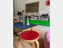 Wohnzimmer mit modernem, farbenfrohem Interieur; im Vordergrund ein Couchtisch mit Dekorationen und ein roter Hocker, im Hintergrund ein Regal mit grünem Schrank, Plattenspieler, Büchern und Dekorationsgegenständen, sowie eine gerahmte, detailreiche Illustration an der Wand.