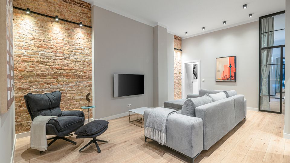 Modernes Wohnzimmer mit freigelegten Backsteinwänden, hellem Holzboden und deckenbündigen Spotleuchten. Ein großes graues Sofa und ein schwarzer Sessel mit Fußhocker stehen vor einer Wand mit Fernseher; daneben führt eine gläserne Schiebetür in einen angrenzenden Raum.