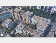 Luftaufnahme eines Wohngebiets mit mehrstöckigen Wohn- und Bürogebäuden im modernen Baustil. Die Gebäude verfügen über Flachdächer, einige mit begrünten Bereichen, und sind von Grünflächen und Gehwegen umgeben. Im Hintergrund sind weitere Wohnhäuser und Bäume sichtbar.