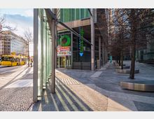 Straßenszene mit modernen Gebäuden und Glasfassaden, im Vordergrund eine Glasüberdachung mit Stützsäulen und gepflastertem Boden. Rechts stehen kahle Bäume mit runden Sitzbänken, links befinden sich ein gelber Linienbus und die Fassade eines Dunkin’ Donuts-Geschäfts.