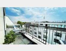 Dachterrasse mit modernen Sitzmöglichkeiten, Pflanzen in Töpfen und Glasgeländer, mit Blick auf umliegende Gebäude und den Berliner Fernsehturm im Hintergrund unter einem bewölkten Himmel.