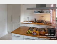 Helle, moderne Küche mit weißen Schränken und Arbeitsplatten aus Holz. In der Küche befinden sich eine Spüle, ein Gasherd, Küchenutensilien, Obstschalen und elektrische Geräte wie Mixer, Kaffeemaschine und Toaster.