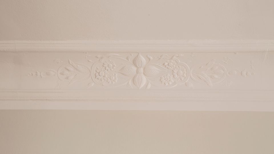 Detailaufnahme einer weißen Zimmerdecke mit breiter Stuckleiste und floralem Reliefmuster. Die glatte Deckenfläche wird von einer horizontal verlaufenden, verzierten Bordüre eingefasst.