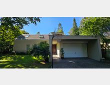 Modernes Einfamilienhaus mit flachem Satteldach, hellbeiger Fassade und Dachfenstern. Links befindet sich ein gepflegter Garten mit Sträuchern, während rechts ein überdachter Bereich mit Garage und Basketballkorb zu sehen ist.